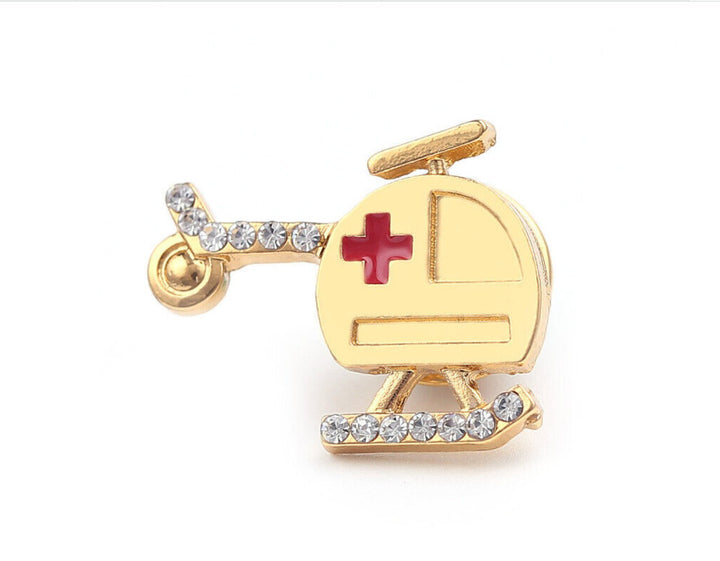 A gold-colored pin in the shape of an air ambulance helicopter, featuring a red cross symbol on the side and adorned with sparkling rhinestones along the base.