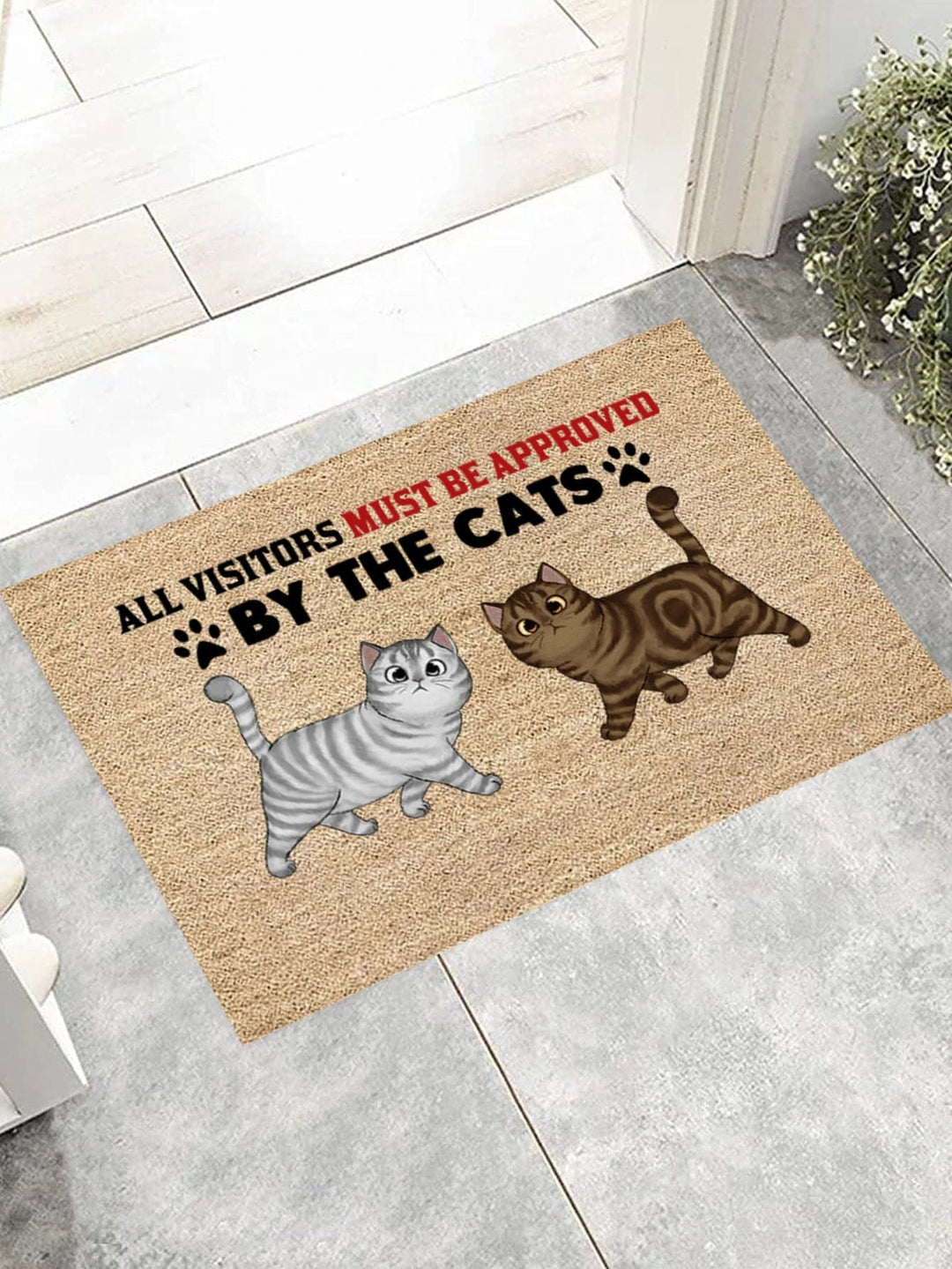 A doormat featuring the text 'ALL VISITORS MUST BE APPROVED BY THE CATS' with illustrations of a gray tabby cat and a brown tabby cat, set against a textured background