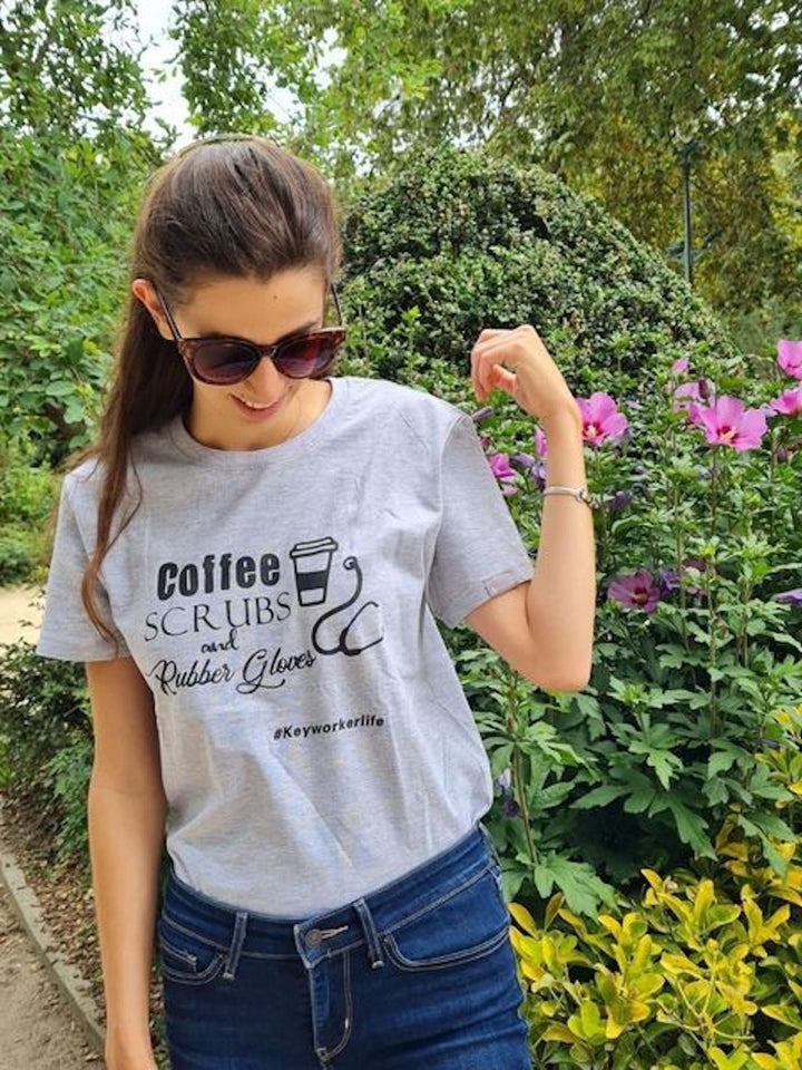 A gray t-shirt featuring the text "Coffee, Scrubs, and Rubber Gloves" along with a graphic of a coffee cup and medical scissors, highlighting the profession of healthcare workers. The shirt is set against a backdrop of greenery and flowers.