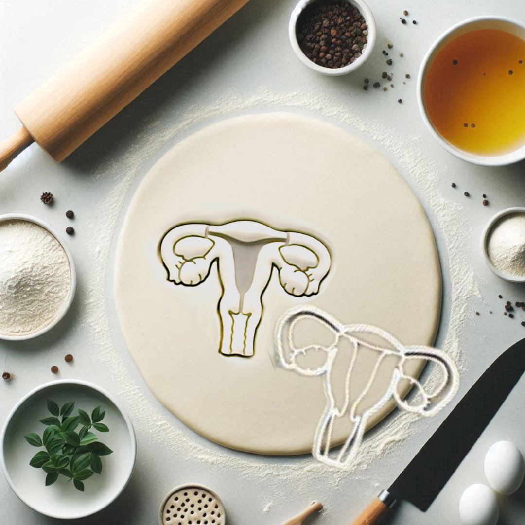 Uterus womb shaped cookie cutter shown imprinted onto rolled out dough. Uterus cookie is next to the rolled out dough. A rolling pin, and baking kitchen items are on the counter around the dough. 